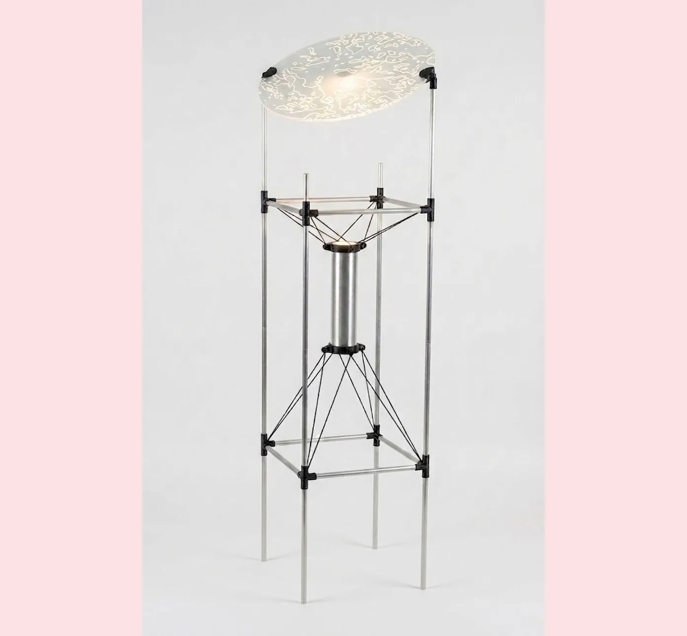 A tall experimental light prototype made from transparent rods, visible tension cables, a central cylindrical element and an illuminated circular surface.