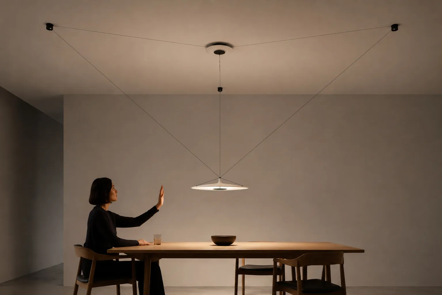A person gestures toward a suspended light positioned above one side of a large dining table.