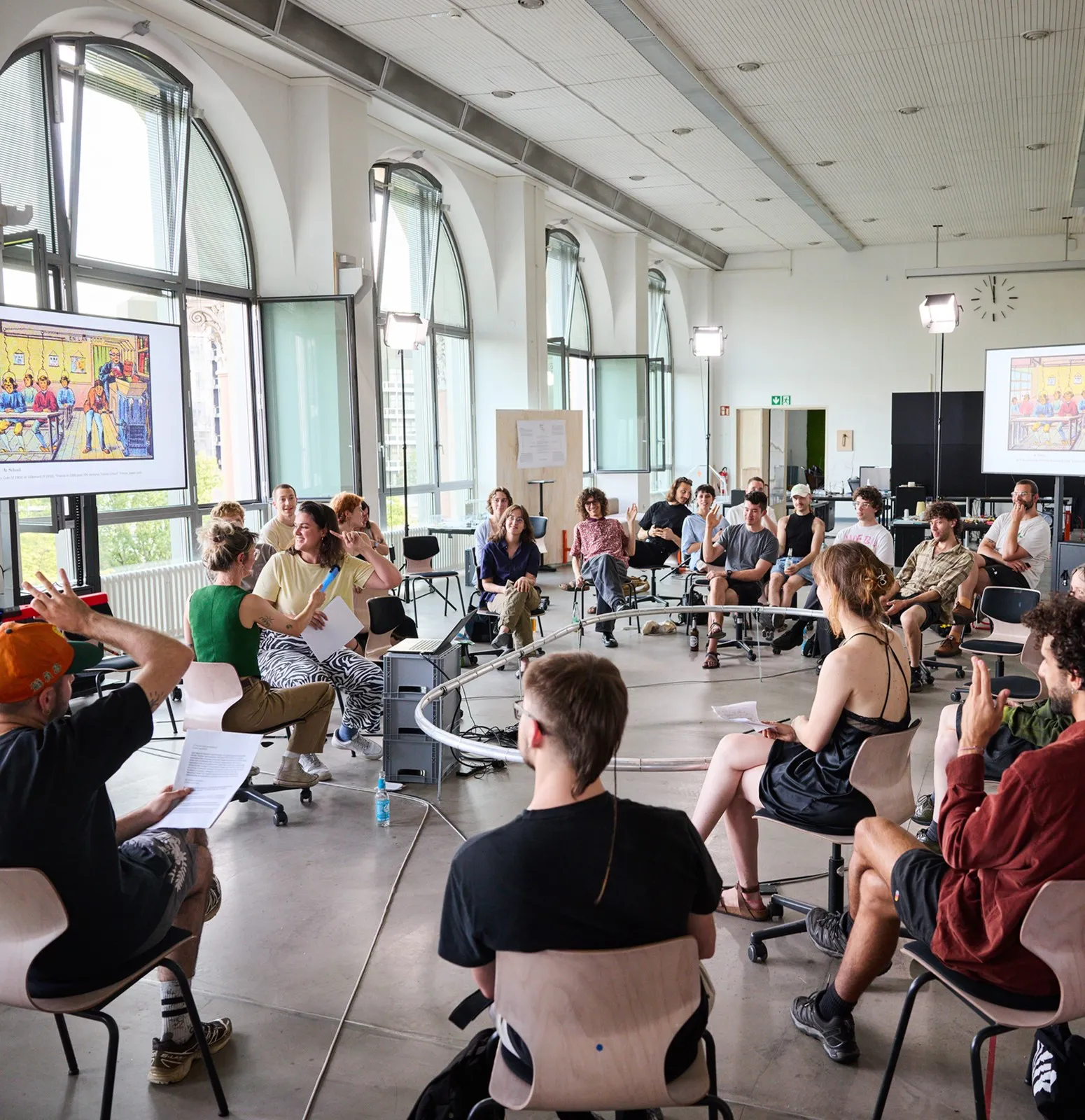 A large discussion circle in the Co-Lab, with participants seated around mobile equipment and presentation screens.